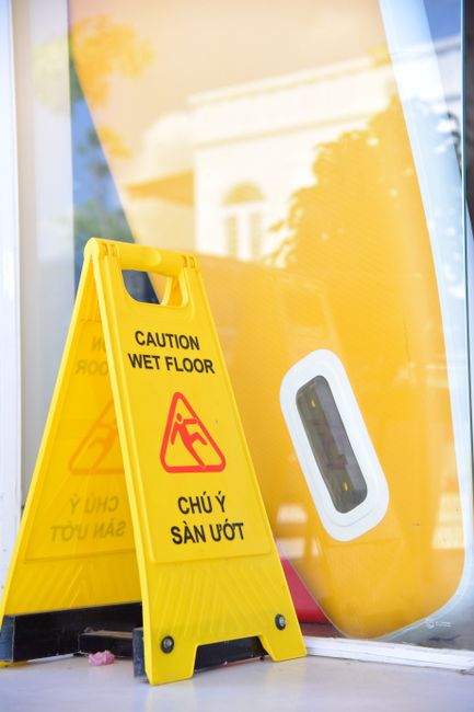 A yellow caution wet floor sign positioned on a smooth, glass door or window with a reflective surface, showing handle and sealing outline, outside a building. The sign has bold black and red text and