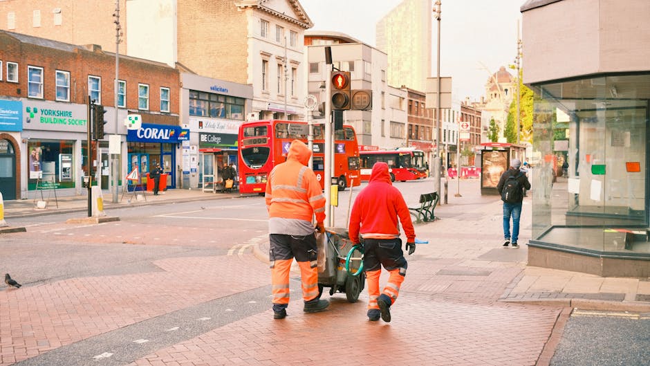 An exterior street scene in Clapham Junction showing a street cleaner operating a green and black commercial cleaning machine on tram tracks, with a few pedestrians walking along the paved sidewalk. T